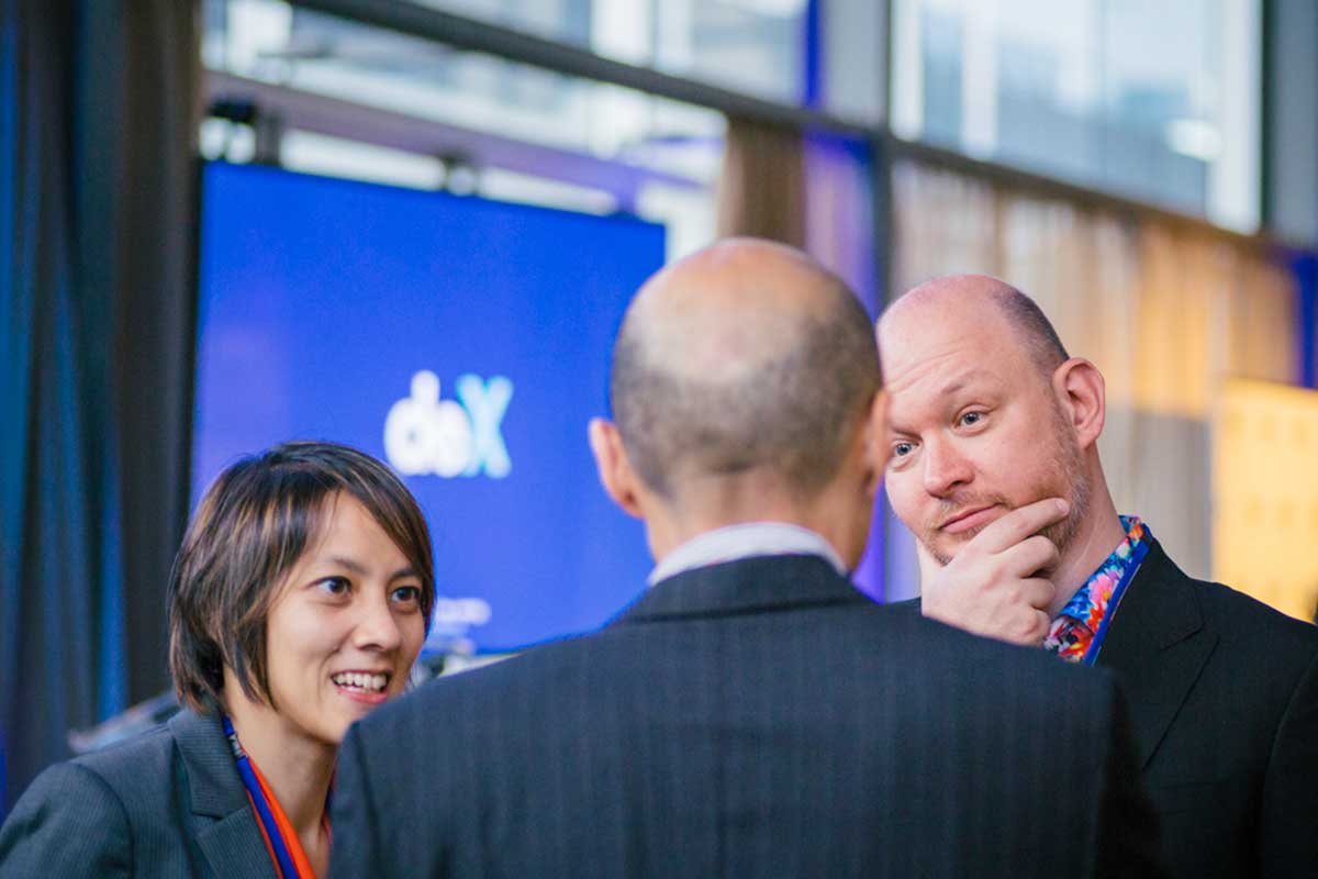 Two men and a woman in suits talking in front of the deX logo